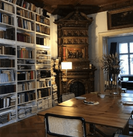 Gemütlicher Raum mit hohen Bücherregalen voller Bücher, einem großen Holztisch mit Stühlen und einem kunstvoll verzierten, historischen Kachelofen; durch das Fenster fällt Tageslicht ein.