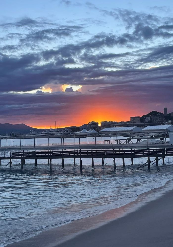 Malerischer Sonnenuntergang über einem ruhigen See oder Meer, mit leuchtenden Orange- und Gelbtönen am Horizont, die sich im Wasser spiegeln. Im Vordergrund ist eine grüne Wiese zu sehen.