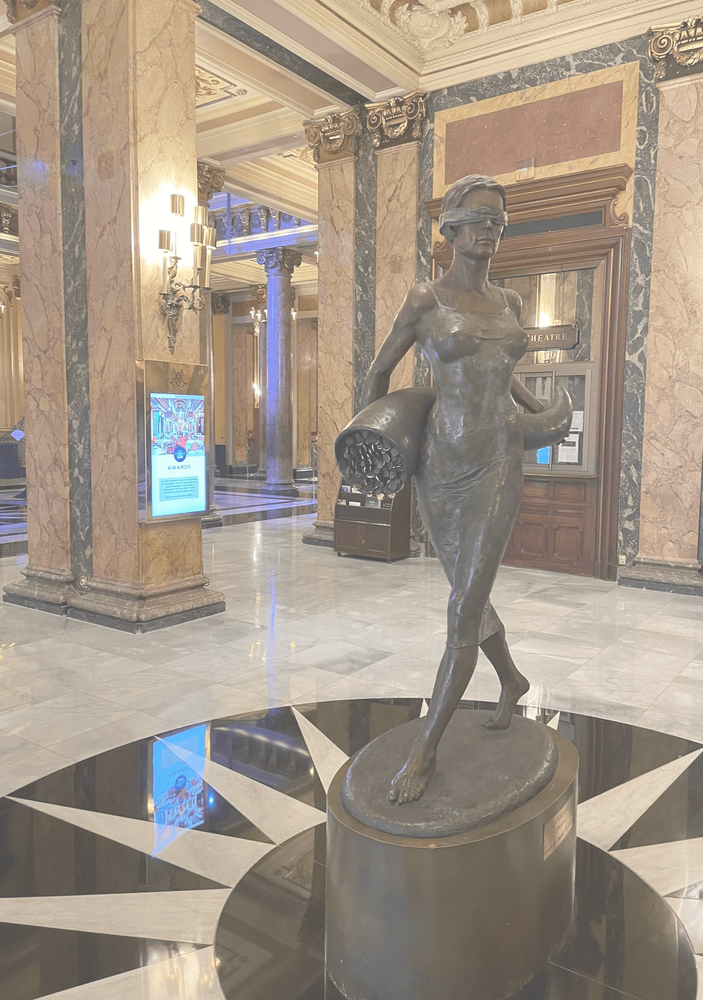 Bronzestatue einer barfüßigen Frau mit verbundenen Augen und einem Füllhorn im Arm, auf einem Podest in einer prächtigen Halle mit Marmorsäulen und kunstvoller Decke.