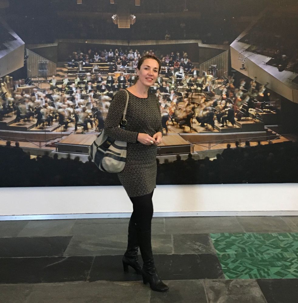 Andrea Berwing steht lächelnd in dunkler Kleidung in einem Raum mit einem großen, verschwommenen Wandgemälde im Hintergrund. Sie hält eine kleine Tasche in der Hand.